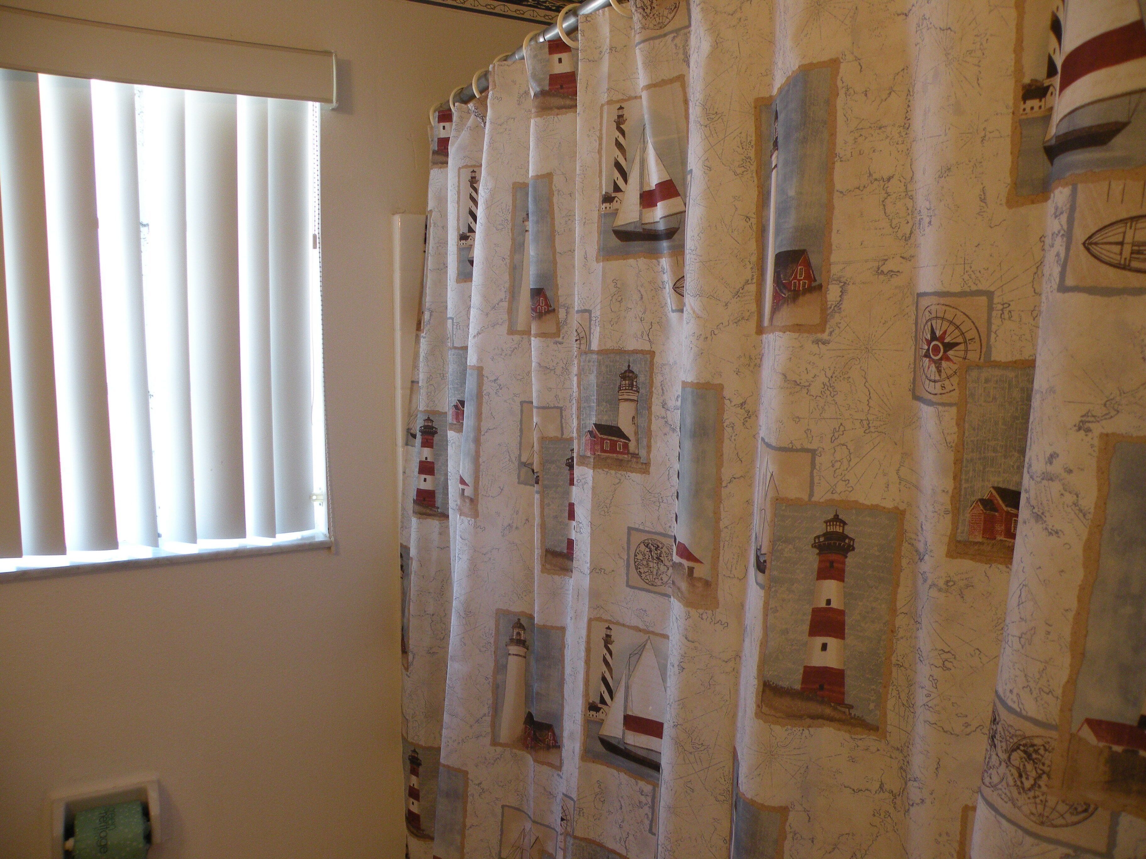 A bathroom with a shower curtain featuring nautical designs, a window with blinds, and a visible electrical outlet.
