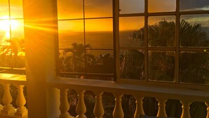 A room with a large window overlooking a view of palm trees and a sunset. -  Sunset Vista (Canefield)