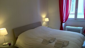 En pænt redt seng med hvide sengetøj og håndklæder, et natbord med en lampe, og et vindue med røde gardiner.