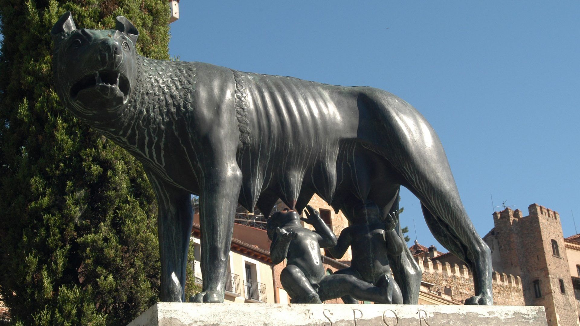 A large bronze statue of a bull with a rider, set against a backdrop of trees and historic buildings.