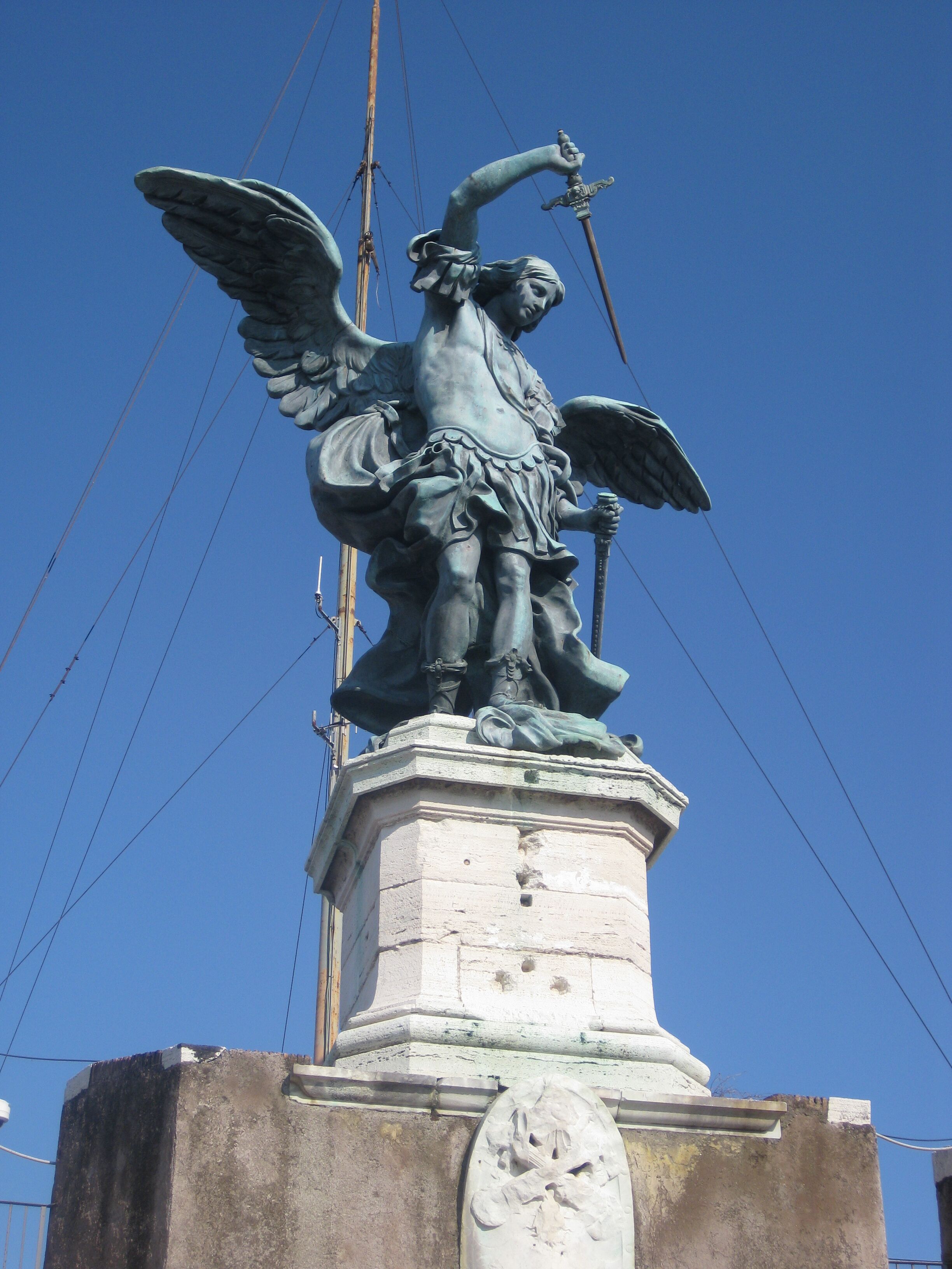 Une statue représentant une figure angélique tenant une lance, placée sur un piédestal dont la base est ornée d’un bas-relief sculpté.