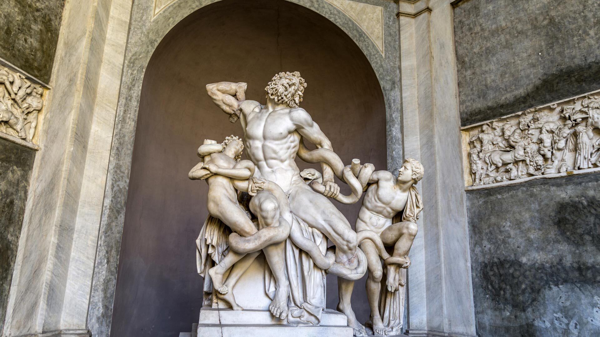 Una scultura in marmo raffigura un uomo muscoloso con un serpente, circondato da figure più piccole, inserita in una nicchia ad arco.