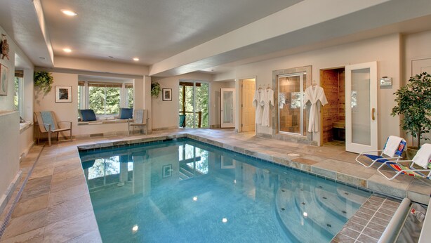 Une piscine intérieure à l’eau d’un bleu limpide, entourée d’un sol et de murs recouverts de carreaux. On y trouve des chaises longues et une porte en bois donnant accès à un vestiaire.