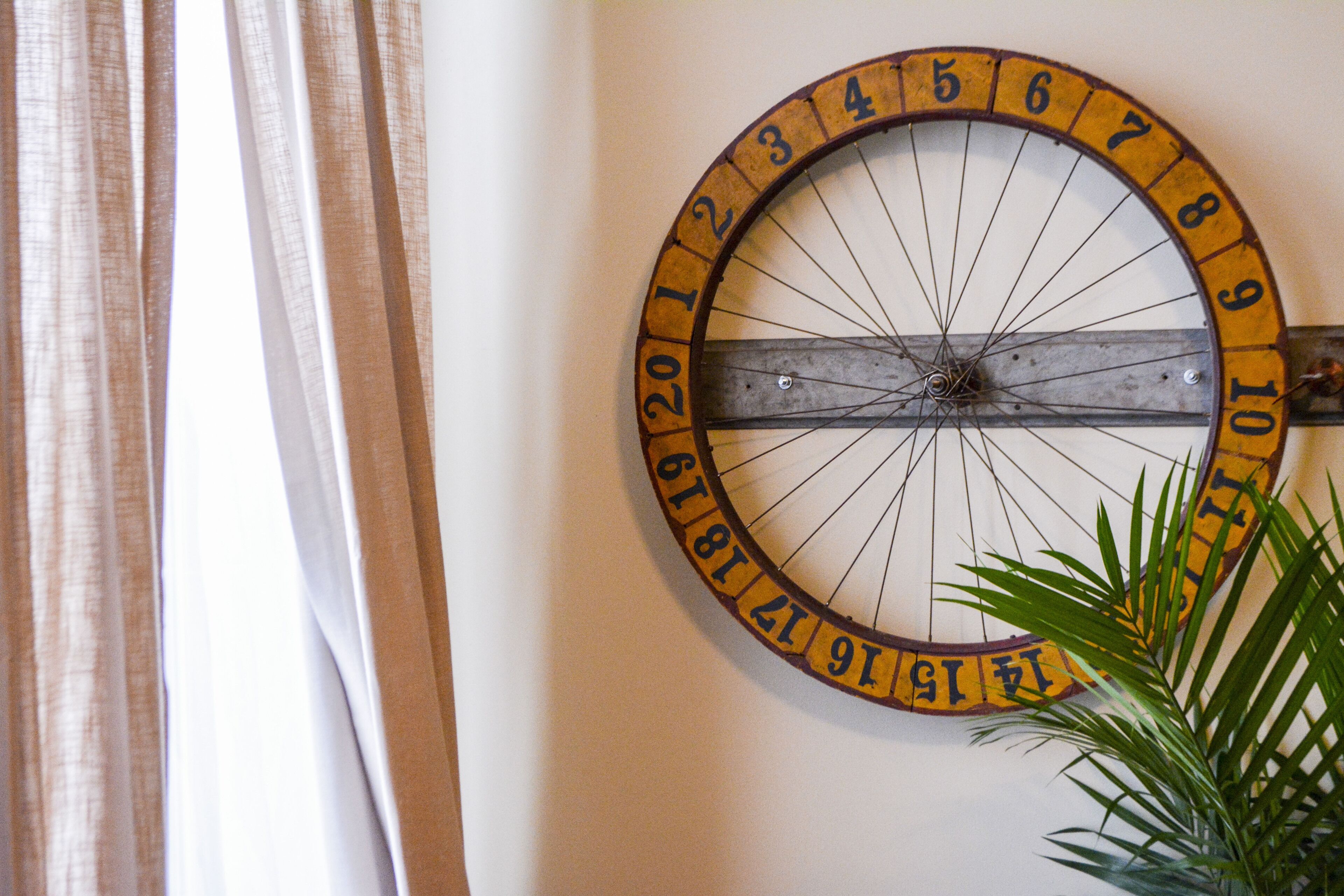 A wall-mounted circular clock with numbers from 1 to 12, mounted on a wall with a curtain partially visible to the left.