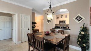 A dining area with a wooden table and chairs, a hanging light fixture, and a kitchen in the background. - Luxury Condo Steps to Disney, Gated Resort Perfect Location, Free Parking & WiFi (Kissimmee)