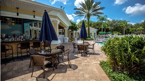 An outdoor bar area with wicker furniture and umbrellas, a pool, and palm trees. - Luxury Condo Steps to Disney, Gated Resort Perfect Location, Free Parking & WiFi (Kissimmee)