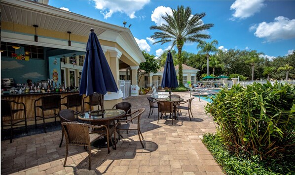 An outdoor bar area with wicker furniture and umbrellas, a pool, and palm trees.