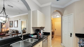 A kitchen with a black countertop, stainless steel sink, and white cabinets. There is a dining area with a table and chairs visible through an archway. - Luxury Condo Steps to Disney, Gated Resort Perfect Location, Free Parking & WiFi (Kissimmee)