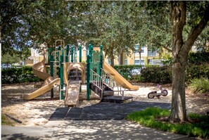 Children can play on the playground's slides and climbing elements in a park-like setting surrounded by trees and a paved path.