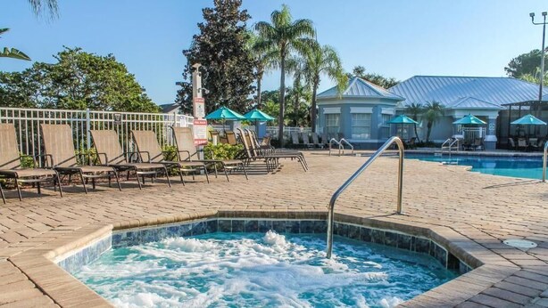 A hot tub surrounded by lounge chairs and umbrellas, with a pool and a building in the background. - Luxury Condo Steps to Disney, Gated Resort Perfect Location, Free Parking & WiFi (Kissimmee)