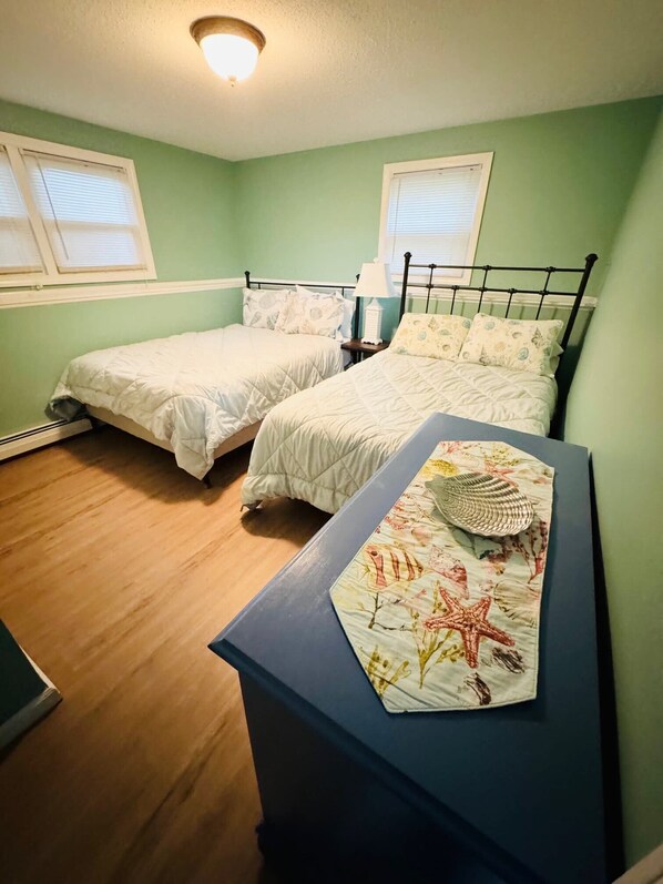 Une chambre avec deux lits, une table de chevet, une commode et une fenêtre avec des stores.