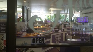 A coffee shop interior with an 'OPEN' sign, a counter displaying pastries and coffee, and a menu board listing coffee types. - Charming Studio in Waikiki - 2 Full size Beds, Free Wi-Fi (Honolulu)