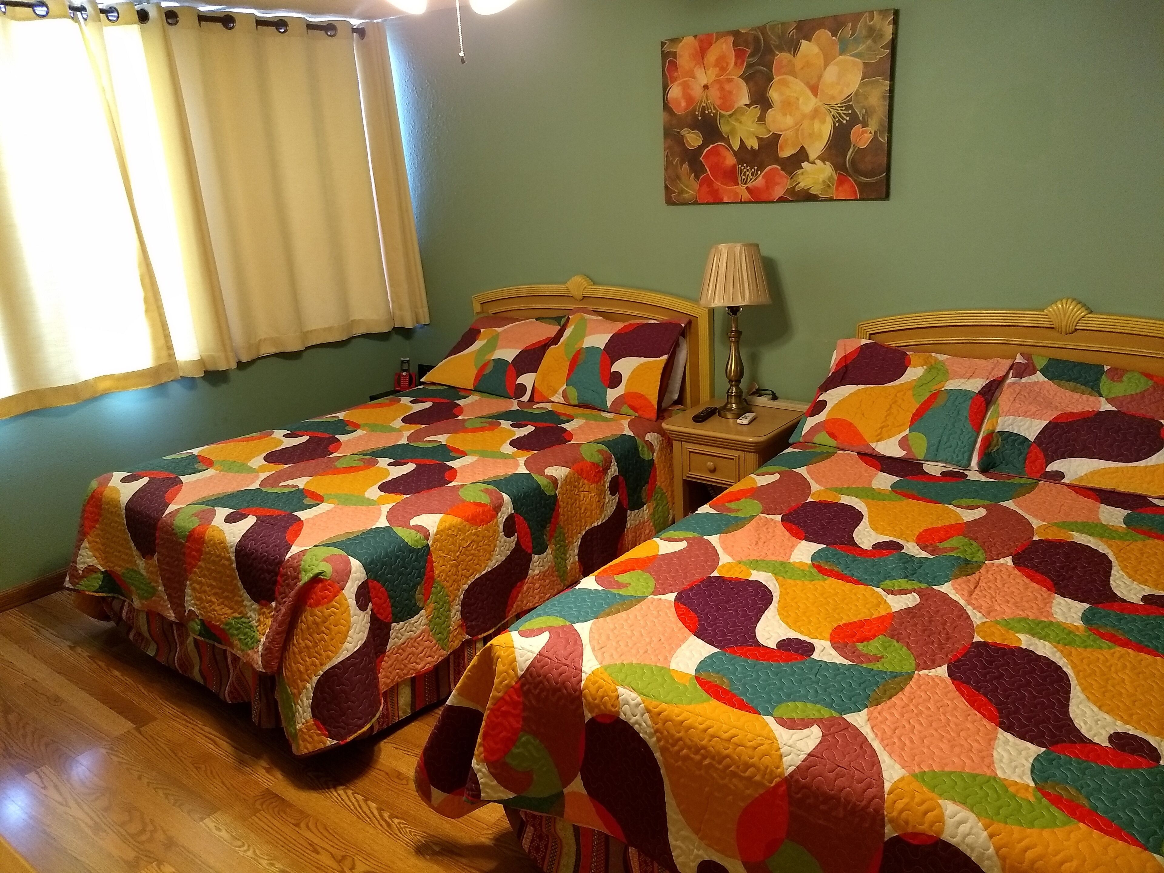 Una habitació amb dos llits, cadascun cobert amb un estrambó de colors, una tauleta de nit amb una làmpada, un quadre a la paret i una finestra amb cortines.