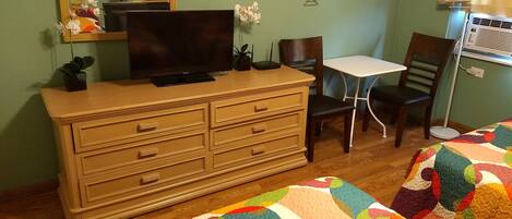 A bedroom with a wooden dresser, a television, a small table, a chair, and a colorful quilt on the bed.