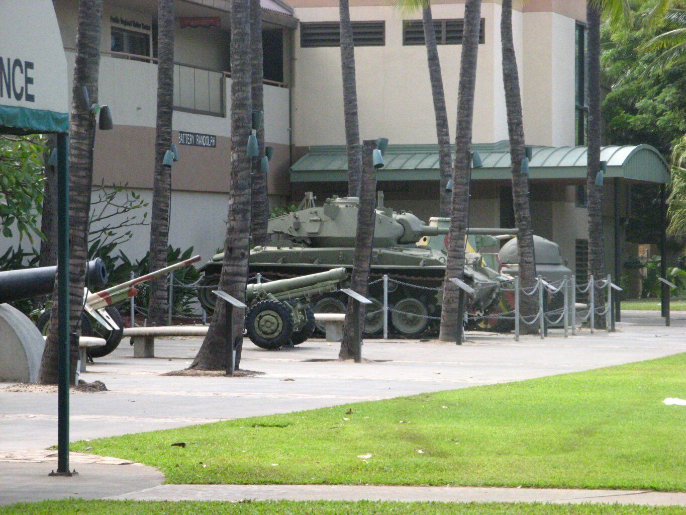 A military vehicle parked in a grassy area with palm trees and a building in the background.