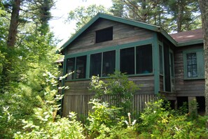A wooden cabin with large windows and a porch, surrounded by dense forest.
