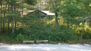A cabin surrounded by dense forest, with a sandy foreground and a wooden bench. - Cottage on Beautiful Long Pond  with Fantastic Sandy Lakefront Beach (Plymouth)