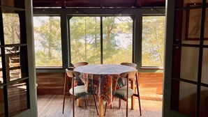 A wooden deck with a round table and chairs, large windows with a view of trees and a body of water. - Cottage on Beautiful Long Pond  with Fantastic Sandy Lakefront Beach (Plymouth)