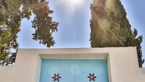 Una puerta azul con dos diseños de flores rojas y un emblema en forma de diamante, sobre un muro blanco con cielo despejado y árboles al fondo.