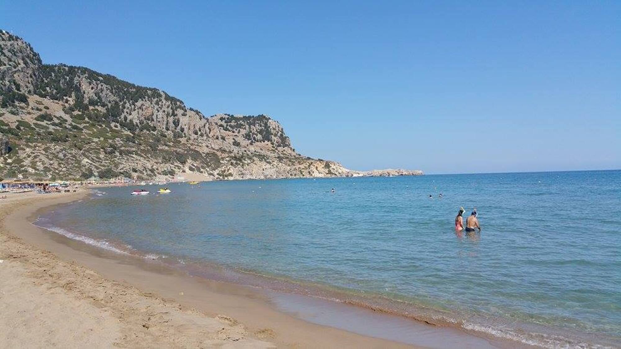 A beach with clear water, a sandy shore, and rocky cliffs in the background.