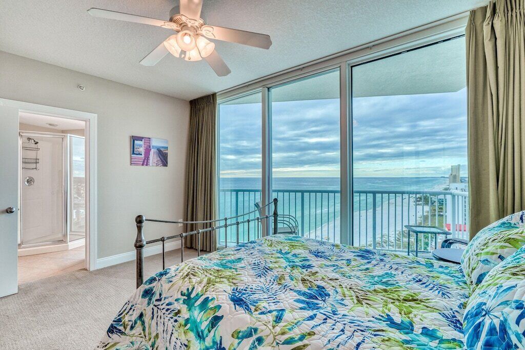 Ein Schlafzimmer mit Bett, Deckenventilator, großem Fenster mit Meerblick und einer Tür zu einem anderen Raum.