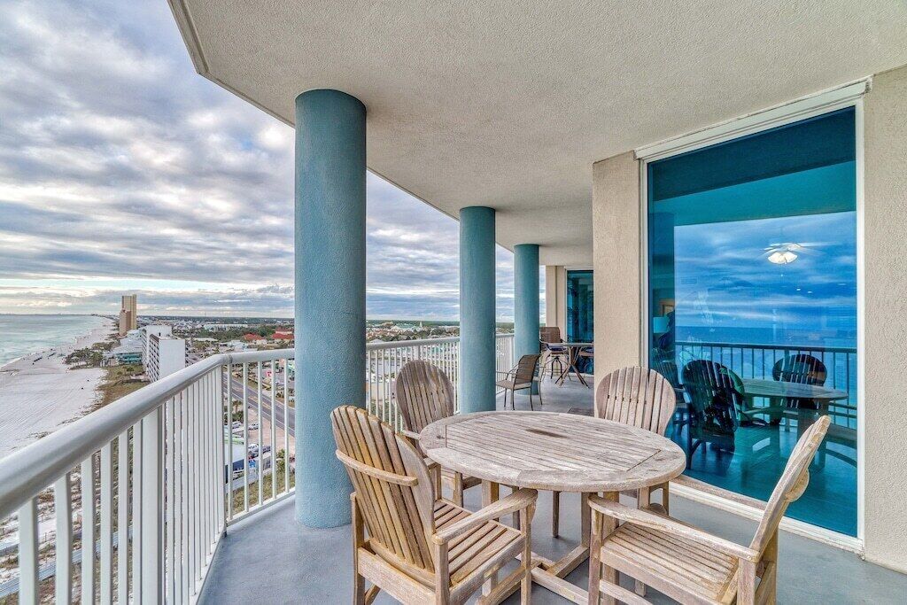 A balcony with a round wooden table and chairs, overlooking a beach and ocean.