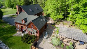 A two-story log cabin with a stone patio and a fenced garden area. - Large Mountain Lodge, Pet Friendly, w/ Hot Tub, and Sports Bar/Game Room. (Ripton)