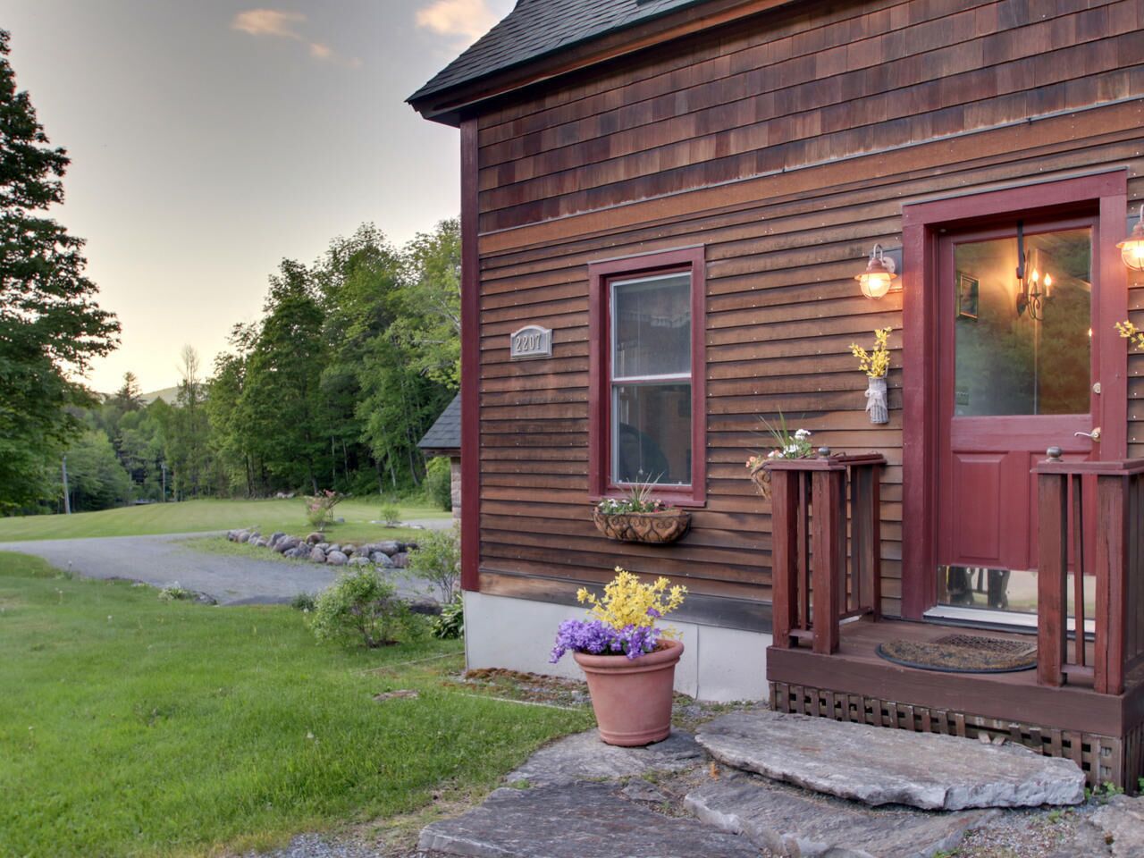 Ett trähus med en röd dörr, ett fönster och en veranda med ett trappsteg av sten. På verandan står blomkrukor och en stensatt gång leder fram till huset.