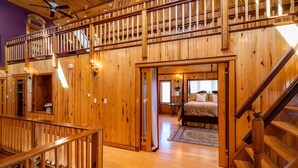 A wooden interior with a double staircase, a bedroom with a bed and a nightstand, and a wooden railing. - Large Mountain Lodge, Pet Friendly, w/ Hot Tub, and Sports Bar/Game Room. (Ripton)