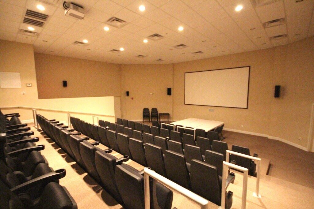 Un salón de conferencias con filas de sillas, una pantalla para proyector y una mesa.
