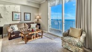 A living room with a sofa, a coffee table, and a chair. There is a window with a view of the sky and a balcony.