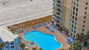 A swimming pool surrounded by lounge chairs and palm trees, adjacent to a building with balconies.