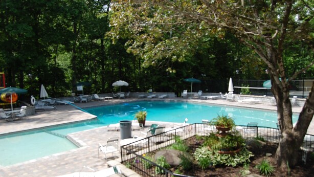 Piscina con sillas de descanso y sombrillas, rodeada de árboles y un jardín cercado.