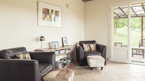 Un salon avec un canapé en cuir, un petit pouf, un bureau avec un ordinateur et des livres, et une grande fenêtre donnant sur l’extérieur.