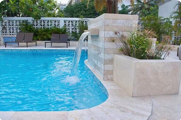 A swimming pool with a waterfall feature, surrounded by a white fence and greenery.