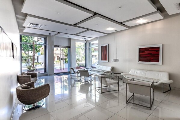 A modern lobby with a white sofa, a white armchair, and a bench. There are large windows and a plant in the corner.