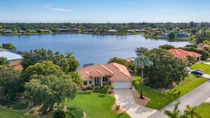 A residential area with a house featuring a red-tiled roof, surrounded by lush greenery and a large body of water. - Beautiful Villa Lake View Heated Pool, Salt💧Spa & Kayak’s🐠 Simply Wonderful! (Cape Coral)