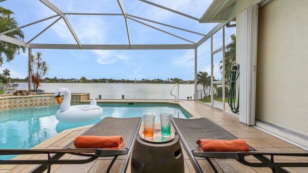 A pool area with lounge chairs, a swan float, and two glasses on a table. - Beautiful Villa Lake View Heated Pool, Salt💧Spa & Kayak’s🐠 Simply Wonderful! (Cape Coral)