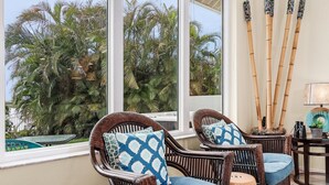 A room with wicker chairs, a small round table, and a view of palm trees through large windows. - Beautiful Villa Lake View Heated Pool, Salt💧Spa & Kayak’s🐠 Simply Wonderful! (Cape Coral)