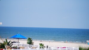 Vista de la playa con una cerca blanca, una sombrilla azul y algunas personas en la arena.