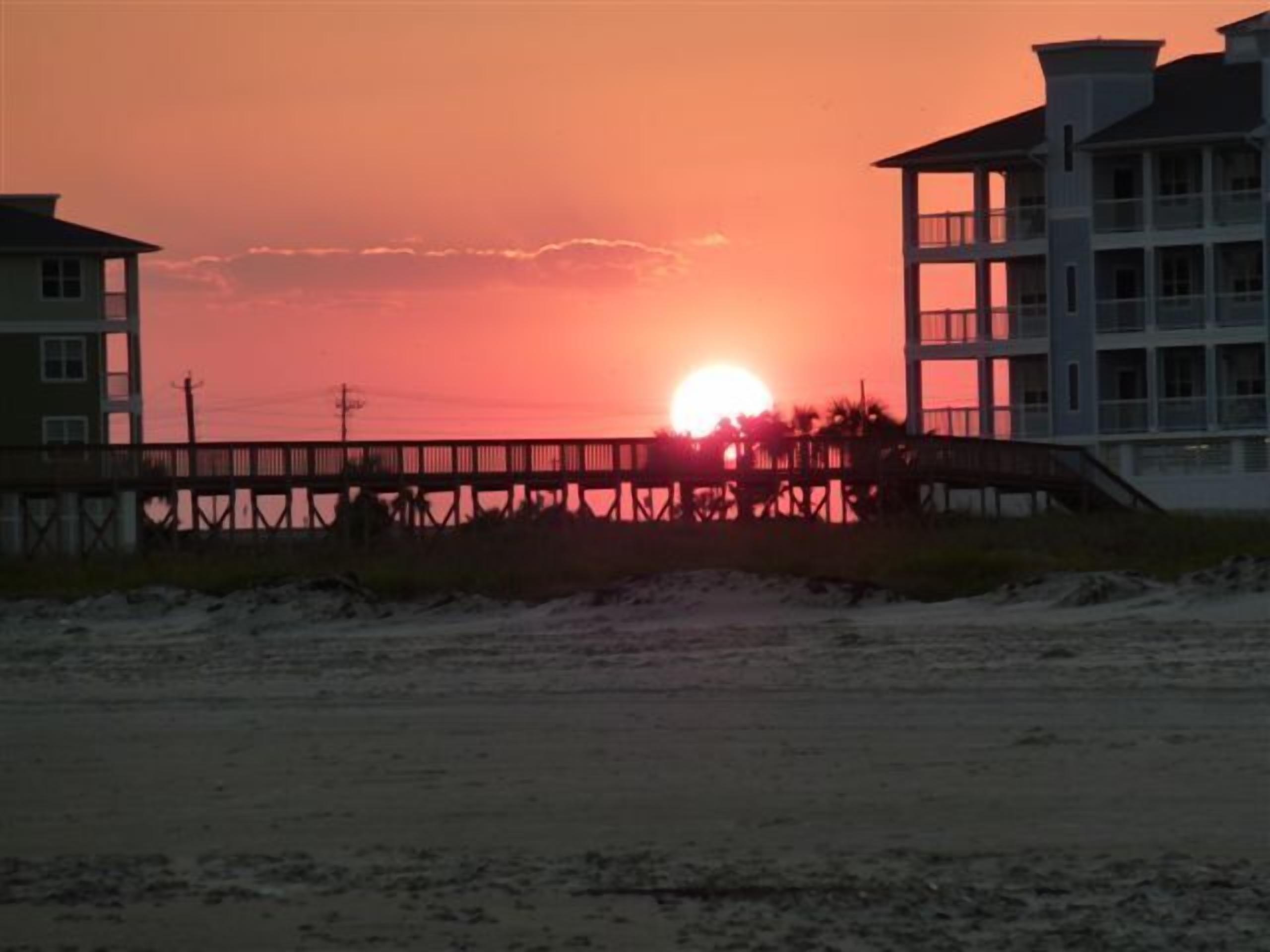 Een zonsondergang boven een strand met een houten pier en gebouwen op de achtergrond.
