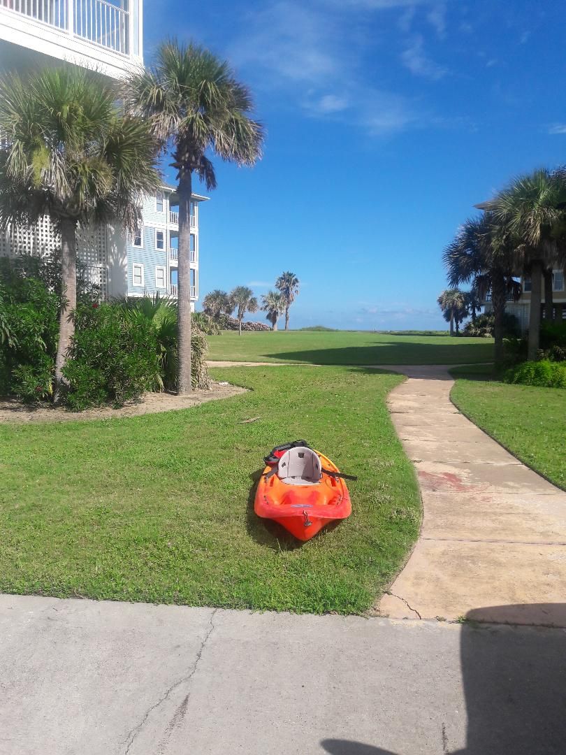 A kayak on a grassy path with a building and palm trees in the background.