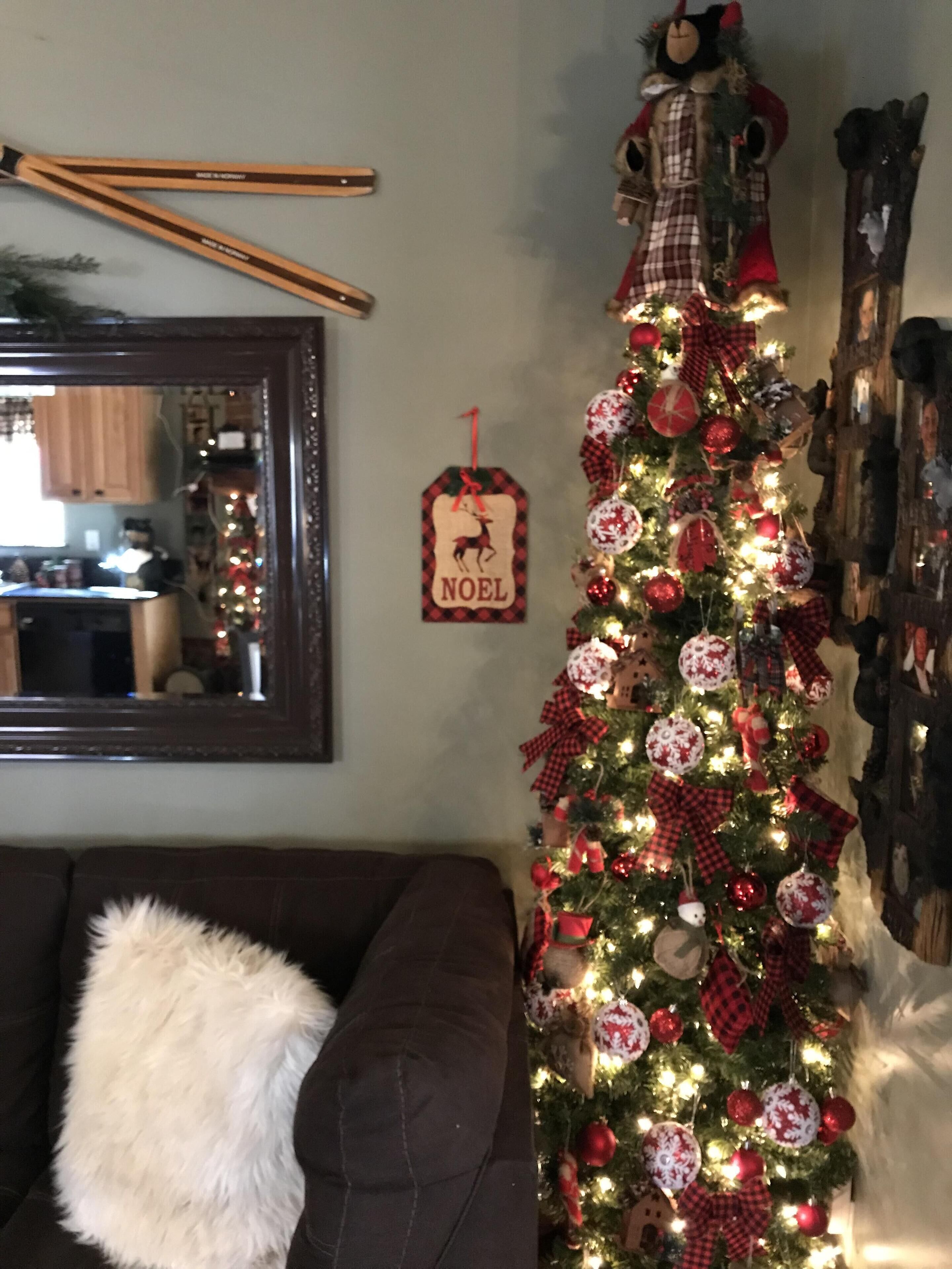 A living room with a Christmas tree decorated with red and white ornaments, a 'NOEL' sign, and a mirror on the wall.