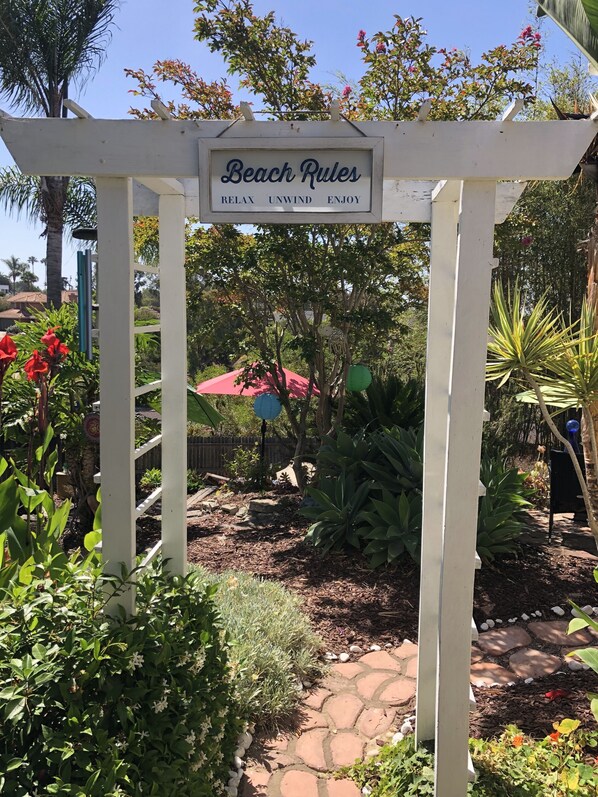 Ein Garten mit einer Pergola, auf der 'Beach Rules' steht, und einem Weg.