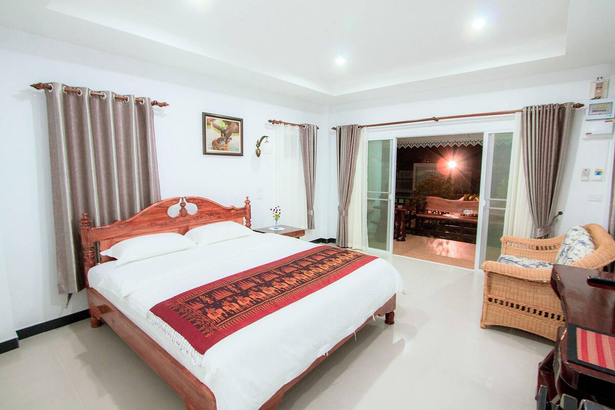 Habitación de hotel con cama de madera, manta estampada roja, una silla de mimbre y vista al área exterior.