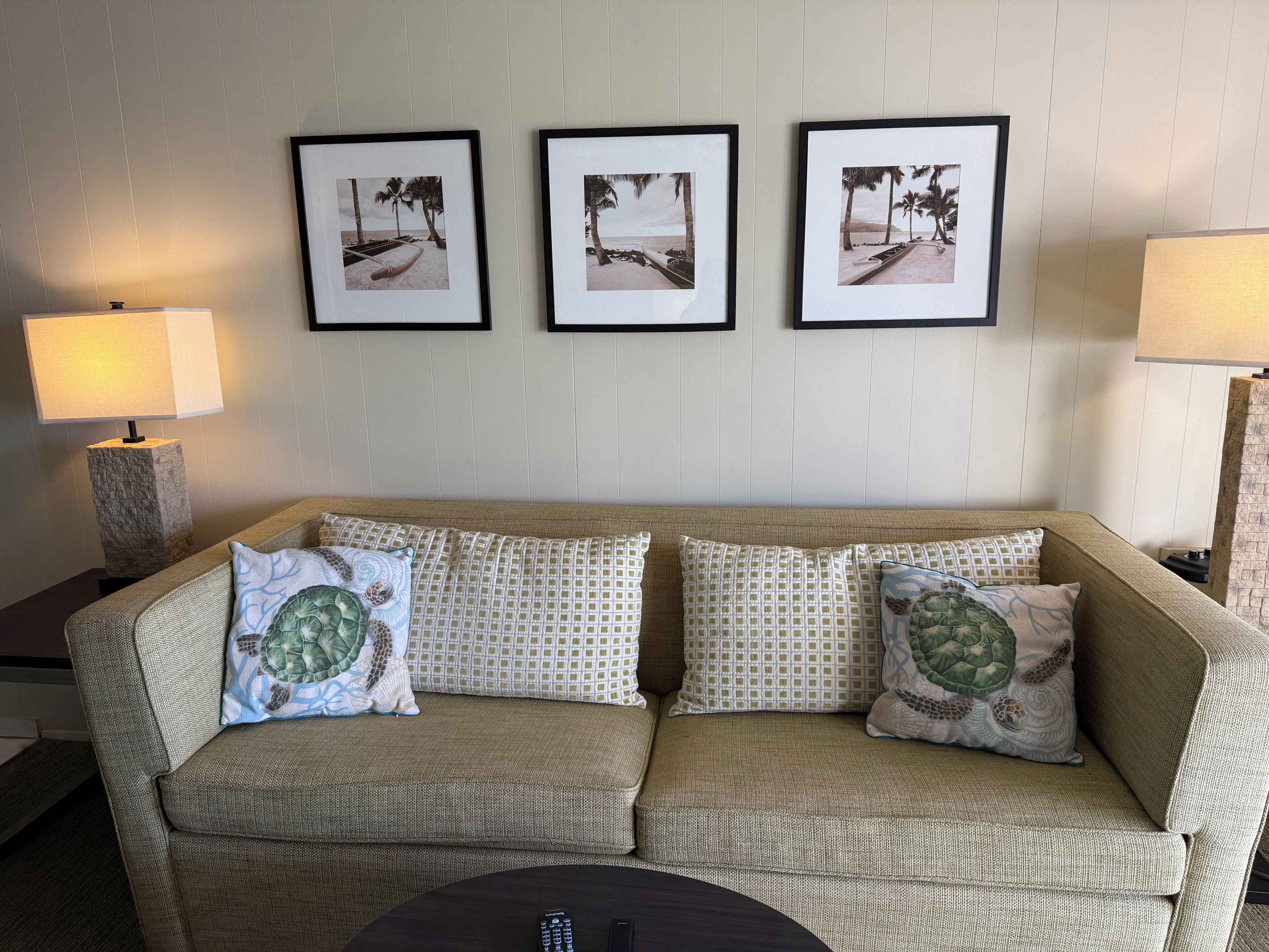 A beige sofa with patterned cushions, a small round table, and three framed beach photographs on the wall.