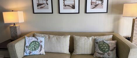 A beige sofa with patterned cushions, a small round table, and three framed beach photographs on the wall.
