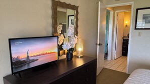A hotel room with a TV displaying a cityscape, a dark wooden dresser, a mirror, and a hallway leading to another room. - Deluxe 1 Bedroom Ocean View Condo 5 minute walk to beach, hot tub and pools  (Kapaa)