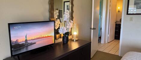 A hotel room with a TV displaying a cityscape, a dark wooden dresser, a mirror, and a hallway leading to another room.
