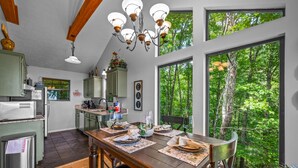 Ein Essbereich mit einem Holztisch, gedeckt für vier Personen, große Fenster mit Blick auf einen Wald und eine Küche im Hintergrund.